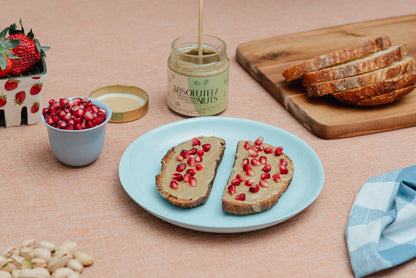 Absolutely Nuts Pistachio Spread with pomegranate seeds on a blue plate, surrounded by a jar of Absolute Nuts, a bowl of pomegranate seeds, and a cutting board with bread.